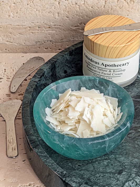White Olivem 1000 emulsifier flakes in a green bowl beside an Amadeus Apothecary facial cream jar.