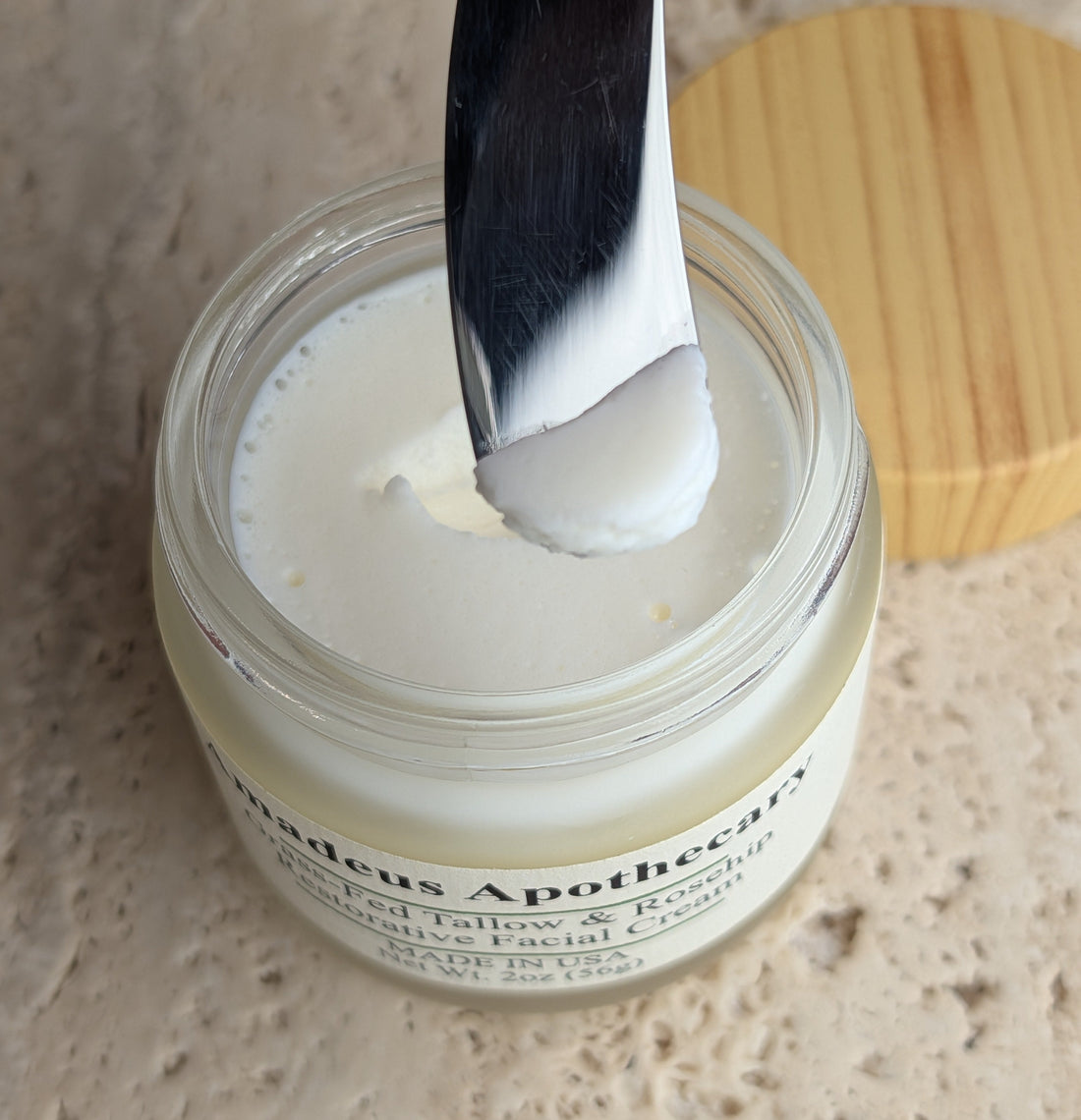 White facial cream on a metal spatula held above an open Amadeus Apothecary jar.
